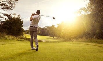 A man swings a golf club on a sunlit golf course, surrounded by lush greenery and trees, with the sun setting in the background, casting a warm glow over the scene.