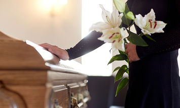 A person dressed in black holds white lilies while touching a wooden coffin in a softly lit room, suggesting a funeral or memorial service.