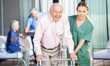 An elderly man using a walker receives assistance from a smiling caregiver in green scrubs, with other seniors and caregivers interacting in a bright, well-lit room in the background.